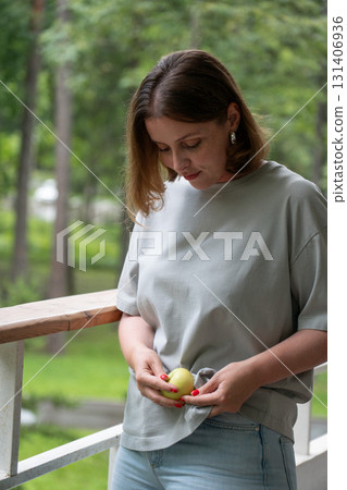 Young woman in light gray t-shirt holding a green apple and looking down thoughtfully while standing on a wooden balcony surrounded by lush greenery 131406936