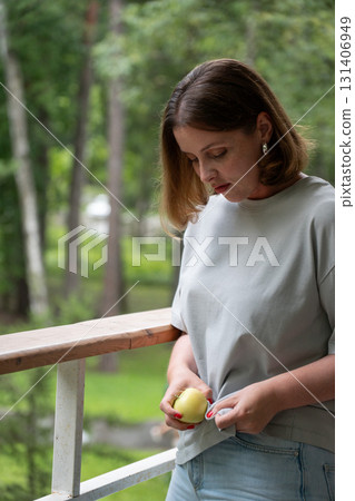 Young woman in light gray t-shirt holding a green apple and looking down thoughtfully while standing on a wooden balcony surrounded by lush greenery 131406949