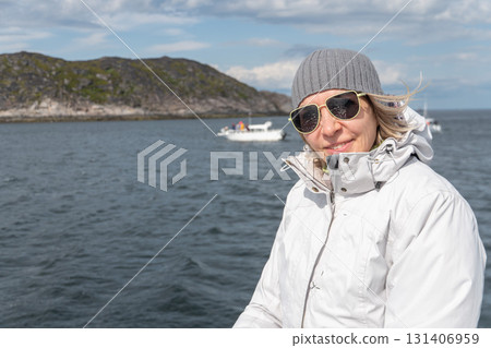 Woman in white jacket and gray knit hat smiles on a boat, rocky coastline and other vessels in the background, clear sky and cold wind 131406959
