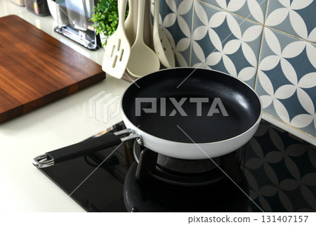 A non-stick frying pan with a white exterior and black handle sits on a black gas stovetop in a modern kitchen 131407157