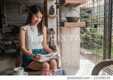 A young woman sits in a stylish cafe, writing in a notebook. The setting features modern decor and abundant natural light. A young woman sits in a stylish cafe, writing in a notebook. The setting features modern decor and abundant natural light. 131408195