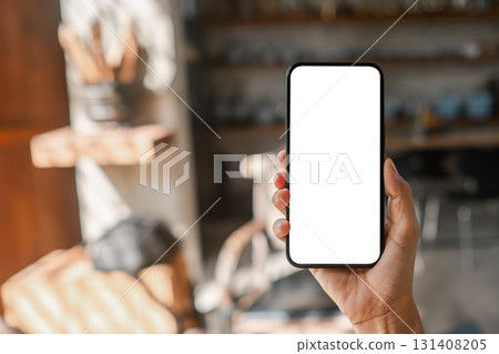 Close-up of a hand holding a smartphone with a blank screen in a cozy cafe, ideal for mockups or design presentations. Close-up of a hand holding a smartphone with a blank screen in a cozy cafe, ideal for mockups or design presentations. 131408205