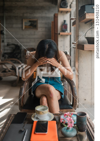 A woman in a modern cafe, holding her head in her hands, with coffee and smartphone on the table, conveying stress or contemplation. 131408210