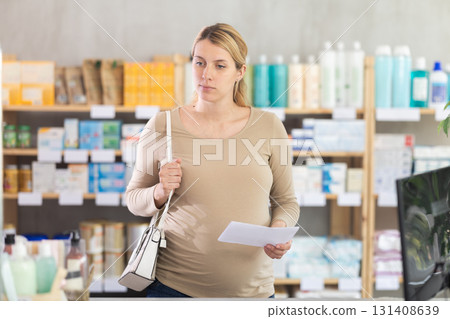 Pregnant woman with doctors prescription is looking for vitamins on pharmacy display case 131408639