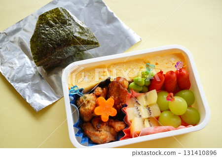 A homey and friendly lunch scene featuring a colorful bento lunch with large rice balls, tamagoyaki (rolled omelet), fried chicken, and other items. A homey and friendly lunch scene featuring a colorful bento lunch with large rice balls, tamagoyaki (rolled omelet), fried chicken, and other items. 131410896