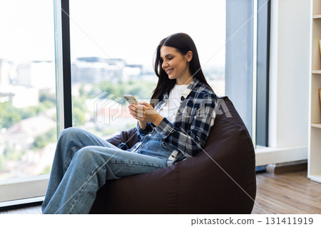 Young adult woman sitting on beanbag chair by window using smartphone. Wearing casual outfit in modern, bright apartment with bookshelves. Relaxed moment of leisure, enjoying serene environment. Young adult woman sitting on beanbag chair by window using smartphone. Wearing casual outfit in modern, bright apartment with bookshelves. Relaxed moment of leisure, enjoying serene environment. 131411919