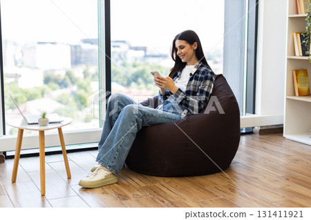 Young adult woman seated on bean bag in modern lounge environment using smartphone. Scene shows casual lifestyle and leisure time indoors. Well-lit surroundings with natural light and wooden flooring. Young adult woman seated on bean bag in modern lounge environment using smartphone. Scene shows casual lifestyle and leisure time indoors. Well-lit surroundings with natural light and wooden flooring. 131411921