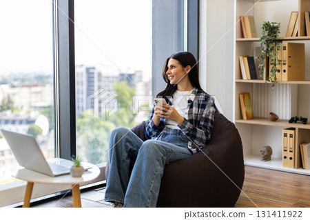 Young adult woman sitting on beanbag chair by window using smartphone. Wearing casual outfit in modern, bright apartment with bookshelves. Relaxed moment of leisure, enjoying serene environment. 131411922