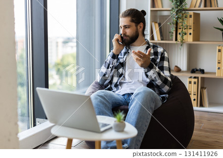 Man in his 30s speaking on phone while working remotely, using laptop and sitting at home 131411926