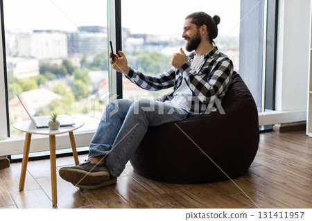Young adult male sitting on beanbag chair in office, chatting through smartphone during workday. Window view in bright modern space portrays casual, multitasking approach to remote communication. Young adult male sitting on beanbag chair in office, chatting through smartphone during workday. Window view in bright modern space portrays casual, multitasking approach to remote communication. 131411957
