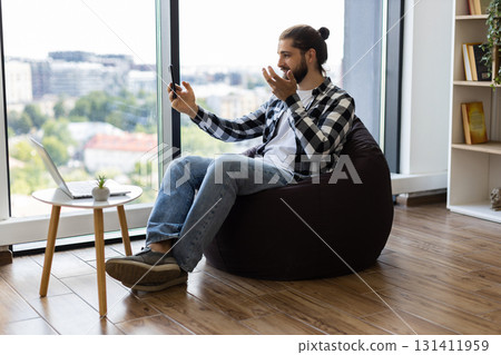 Young adult male sitting in cozy room using smartphone for video call communication 131411959