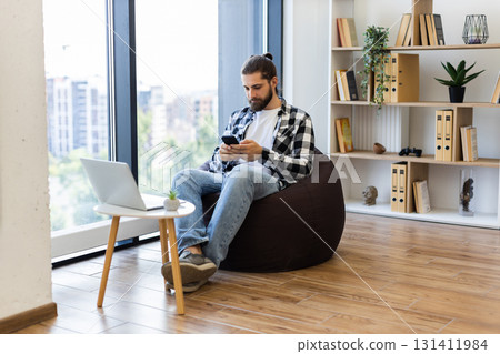 Young adult male using smartphone sitting on beanbag in bright room with minimalist decor, large bookshelves, and laptop table, displaying technology, communication, and casual workspace concepts. Young adult male using smartphone sitting on beanbag in bright room with minimalist decor, large bookshelves, and laptop table, displaying technology, communication, and casual workspace concepts. 131411984