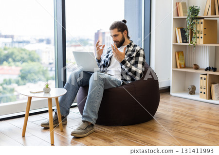 Young adult male wearing casual flannel and jeans using laptop in contemporary office space featuring modern decor and bright sunlight from glass windows Young adult male wearing casual flannel and jeans using laptop in contemporary office space featuring modern decor and bright sunlight from glass windows 131411993
