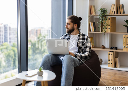 Young adult male with beard and long hair using laptop in cozy home setting. Relaxed atmosphere, comfortable seating, natural light, casual outfit, productive moment, contemporary interior design Young adult male with beard and long hair using laptop in cozy home setting. Relaxed atmosphere, comfortable seating, natural light, casual outfit, productive moment, contemporary interior design 131412005
