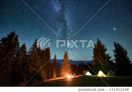 Panoramic view of grassy hill with illuminated camp tents and coniferous trees under starry sky. Night camping in mountains under beautiful sky with stars. Concept of hiking, nighttime and galaxy. Panoramic view of grassy hill with illuminated camp tents and coniferous trees under starry sky. Night camping in mountains under beautiful sky with stars. Concept of hiking, nighttime and galaxy. 131412240