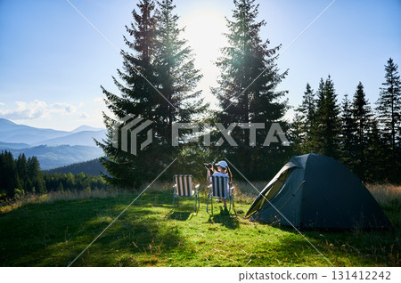 Woman camper relaxes next to tent with photo camera. Female tourist surrounded by tall evergreen trees and bathed in soft evening light, capturing peacefulness of moment. Woman camper relaxes next to tent with photo camera. Female tourist surrounded by tall evergreen trees and bathed in soft evening light, capturing peacefulness of moment. 131412242