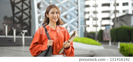 Smiling businesswoman carrying documents, standing outside a modern building Smiling businesswoman carrying documents, standing outside a modern building 131416192