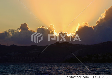 Dramatic sunset or sunrise over a dark mountain silhouette and sea, with golden sun rays bursting through the heavy clouds, creating a majestic and awe inspiring scene. 131417180