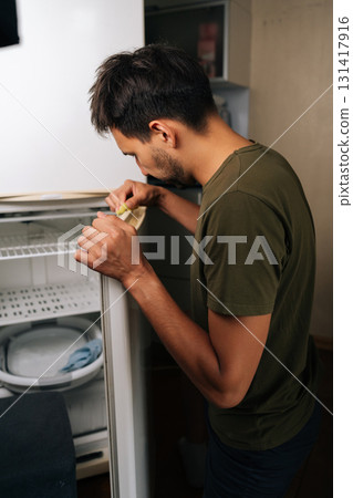 Vertical shot of focused young man cleaning refrigerator door seal with yellow sponge, focusing on detailed sanitation and preserving appliance functionality during routine kitchen maintenance. Vertical shot of focused young man cleaning refrigerator door seal with yellow sponge, focusing on detailed sanitation and preserving appliance functionality during routine kitchen maintenance. 131417916