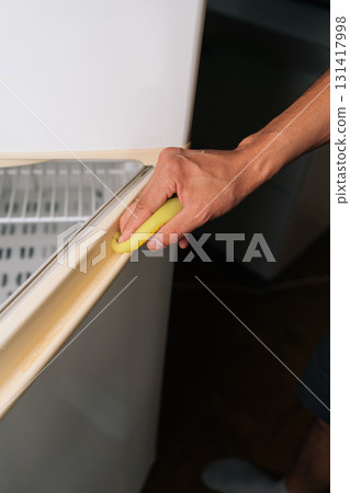 Close-up vertical shot of unrecognizable person cleaning refrigerator door seal with sponge, focusing on detailed sanitation and preserving appliance functionality during routine kitchen maintenance. 131417998