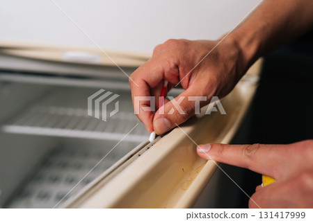 Close-up hands of unrecognizable man cleaning rubber seal of refrigerator door using cotton swab, ensuring hygiene and proper sealing. Concept of domestic work, housekeeping and household. 131417999