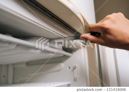Detail cropped shot of unrecognizable man using knife to scraping away thick ice buildup from inside refrigerator, emphasizing routine task of maintaining clean kitchen appliance, close-up. 131418006