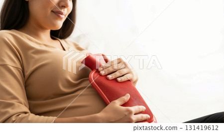Woman holding a red hot water bottle on her abdomen for comfort and pain relief. Self-care for menstrual cramps and warmth. Copy space 131419612