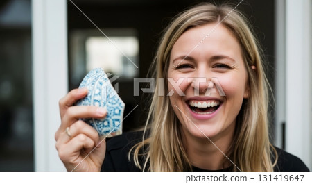 Happy blonde woman laughing while holding a broken piece of a patterned ceramic. Authentic portrait of genuine joy and amusement 131419647