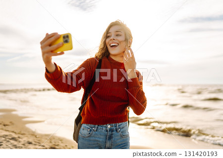 Young woman joyfully taking a selfie on a sunny beach during sunset with ocean waves 131420193