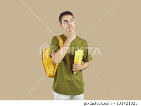 Young thinking brunette student boy holding a textbook with backpack on studio beige background. Young thinking brunette student boy holding a textbook with backpack on studio beige background. 131421023