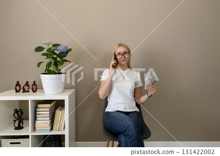 Young woman holding money and talking on phone at home 131422002