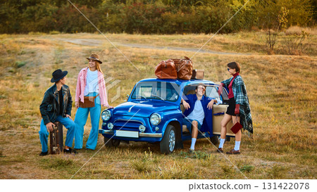 Group of friends in retro style outfits standing by blue car with luggage on roof 131422078