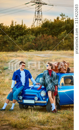 Stylish young couple leaning on retro blue car with luggage on roof smiling in field Stylish young couple leaning on retro blue car with luggage on roof smiling in field 131422083