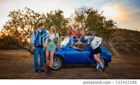 Group of young friends with luggage standing by blue retro car at sunset 131422108