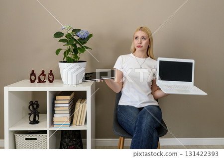 Woman comparing tablet and laptop with unsure expression Woman comparing tablet and laptop with unsure expression 131422393