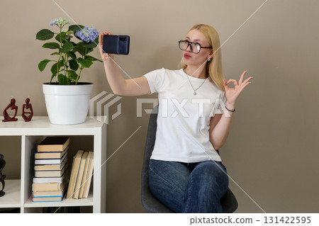 Woman taking selfie showing OK hand gesture in cozy home setting Woman taking selfie showing OK hand gesture in cozy home setting 131422595