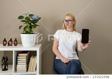 Woman showing smartphone screen while sitting in modern home interior Woman showing smartphone screen while sitting in modern home interior 131422596