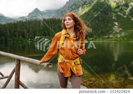 Female traveler with hiking backpack poses against backdrop of mountain lake. Adventure concept. 131422924