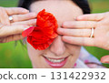 A woman playfully hides her eye with a vibrant red poppy flower, smiling gently 131422932