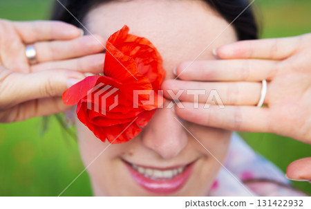 A woman playfully hides her eye with a vibrant red poppy flower, smiling gently 131422932