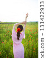 A woman in a pink floral dress stands in a poppy field, arms raised, enjoying the summer sun 131422933