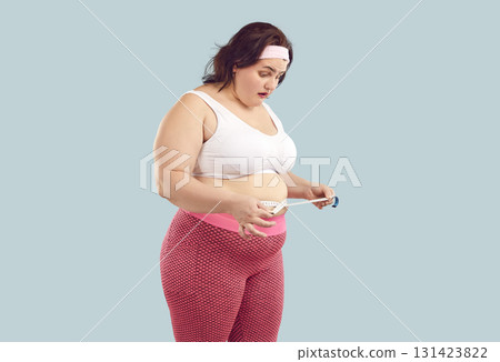 Overweight woman with fat belly looks at measuring tape with surprised face expression 131423822