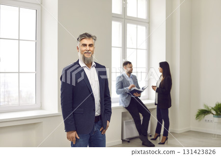 Smiling mature businessman standing in office with colleagues on background Smiling mature businessman standing in office with colleagues on background 131423826