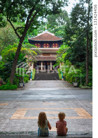 Two children sit on steps facing a traditional Vietnamese temple surrounded by lush tropical greenery in Saigon. Peaceful moment in cultural setting, perfect for family travel, and lifestyle themes. Two children sit on steps facing a traditional Vietnamese temple surrounded by lush tropical greenery in Saigon. Peaceful moment in cultural setting, perfect for family travel, and lifestyle themes. 131424456
