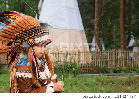 Portrait of young girl wearing a traditional Indian feathered costume. The image was taken outdoors in a forest like setting, evoking a cultural or educational experience for children. Portrait of young girl wearing a traditional Indian feathered costume. The image was taken outdoors in a forest like setting, evoking a cultural or educational experience for children. 131424487