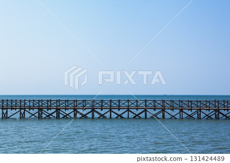 Long wooden bridge stretches across calm turquoise water. Traditional Indonesian design, serene seascape, clear blue sky create peaceful tropical atmosphere, ideal for travel and tourism promotions. Long wooden bridge stretches across calm turquoise water. Traditional Indonesian design, serene seascape, clear blue sky create peaceful tropical atmosphere, ideal for travel and tourism promotions. 131424489