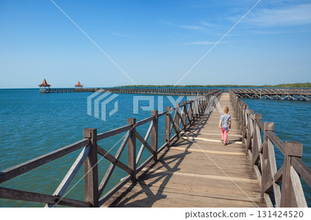 Little boy walks along a long wooden pier stretching into the blue sea on sunny day. Clear blue sky, calm ocean create peaceful, tropical atmosphere. Travel with kids, summer, beach vacation Little boy walks along a long wooden pier stretching into the blue sea on sunny day. Clear blue sky, calm ocean create peaceful, tropical atmosphere. Travel with kids, summer, beach vacation 131424520