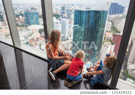Woman, two children look at panoramic view of Ho Chi Minh City from modern skyscrapers observation deck. Urban lifestyle, travel with kids, and high-rise cityscape concept with Southeast Asian charm. 131424521