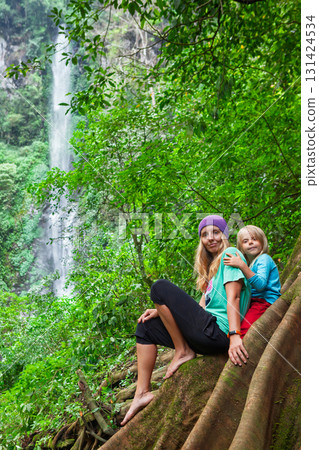 Mother and child rest on giant tree roots in lush tropical jungle near high waterfall. Perfect for eco-tourism, nature escapes, family friendly hikes with breathtaking views and peaceful surroundings. 131424534