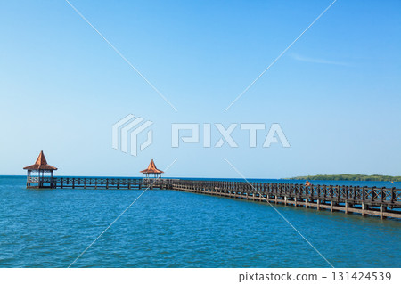 People walk by long wooden pier with traditional gazebos stretches over calm turquoise sea under clear blue sky. Perfect tropical scenery for travel, resort, summer vacation, coastal tourism themes. People walk by long wooden pier with traditional gazebos stretches over calm turquoise sea under clear blue sky. Perfect tropical scenery for travel, resort, summer vacation, coastal tourism themes. 131424539